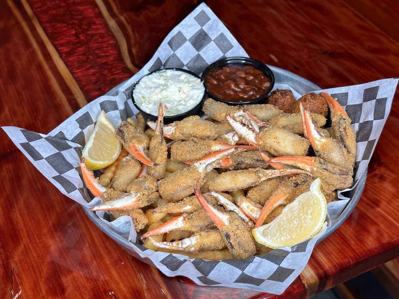 A basket of golden fried crab claws with fries, slaw, baked beans and hush puppies on checkered paper